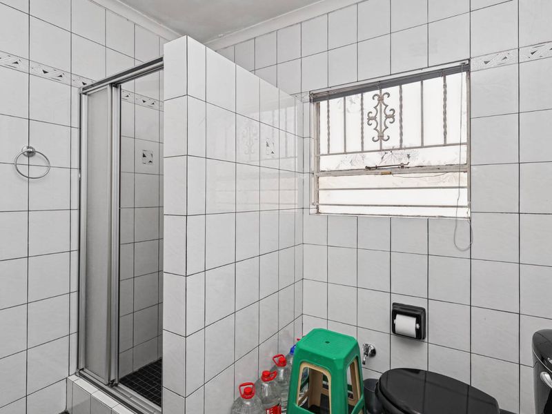 A functional bathroom featuring a shower enclosure, tiled walls, and a window with security bars, requiring some cosmetic updates to improve its overall condition.
