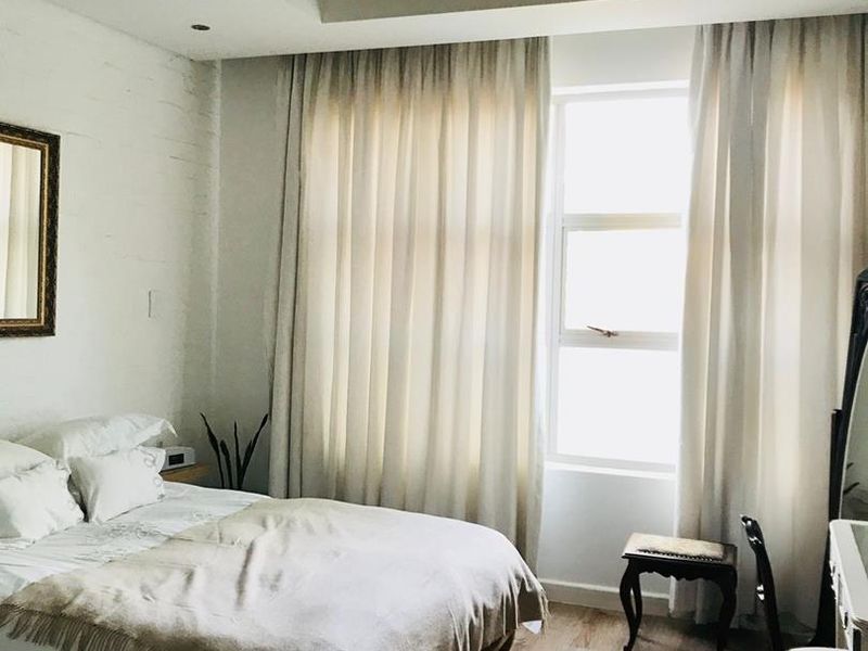 A bright bedroom featuring an exposed concrete ceiling, laminate flooring, and large windows with floor-to-ceiling curtains.