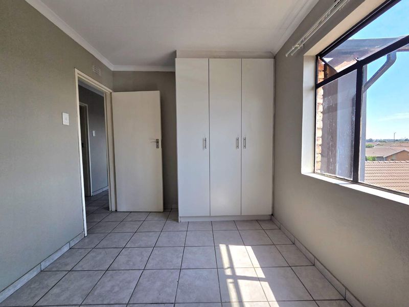 A bright, empty bedroom featuring neutral tiled flooring, a built-in wardrobe, and a large window providing natural light and an exterior view.