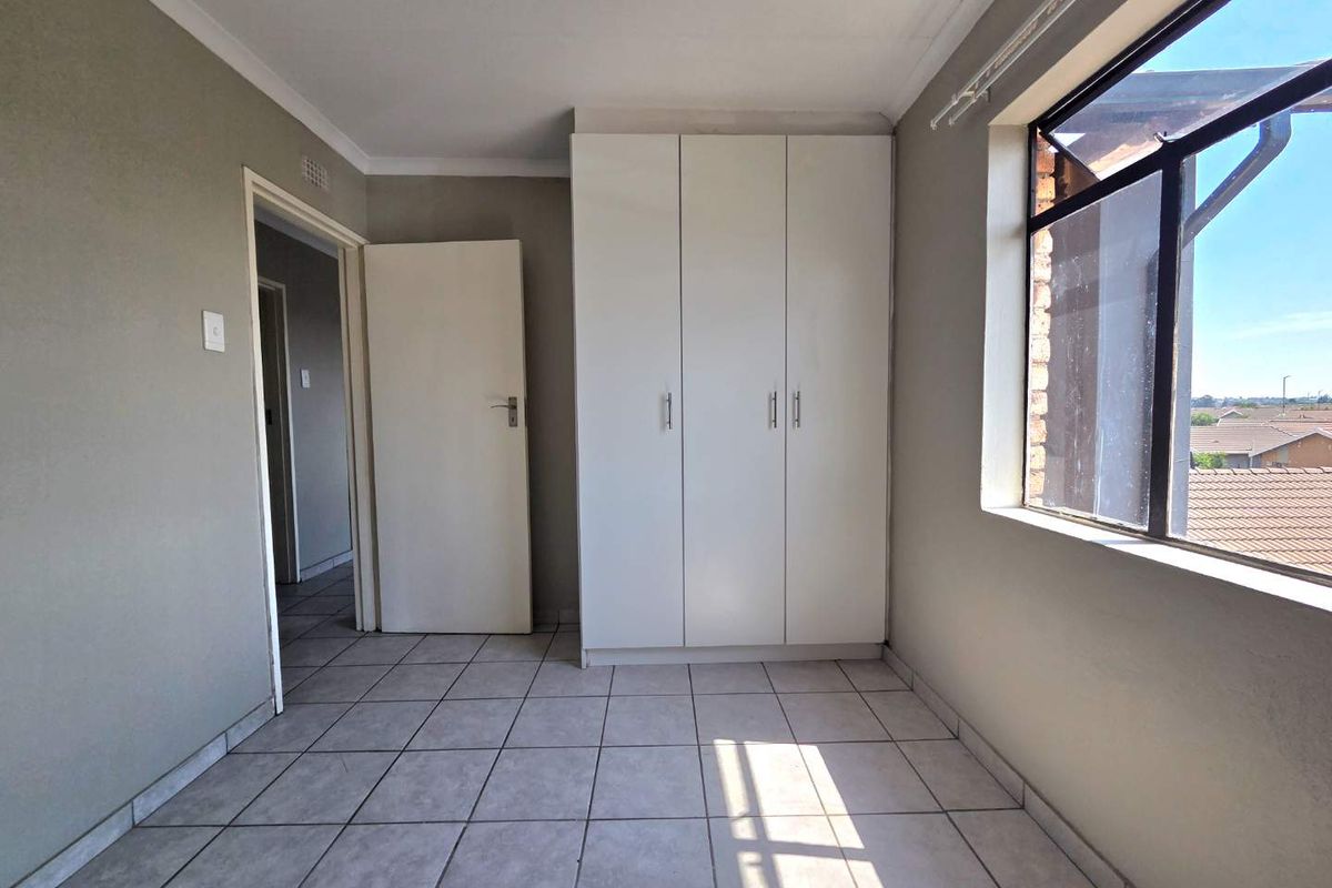 A bright, empty bedroom featuring neutral tiled flooring, a built-in wardrobe, and a large window providing natural light and an exterior view.