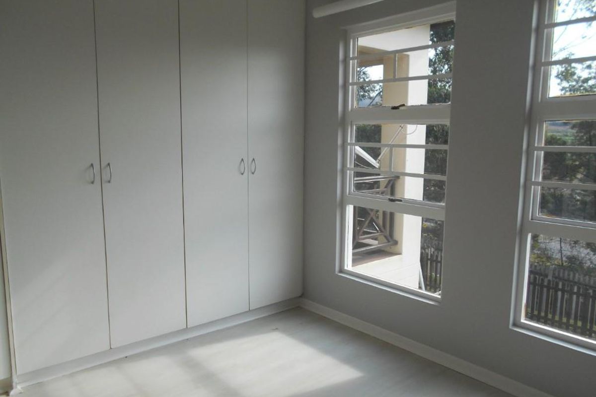 A bright, empty bedroom featuring neutral walls, light-toned flooring, and ample built-in cupboard storage with large windows fitted with security bars.