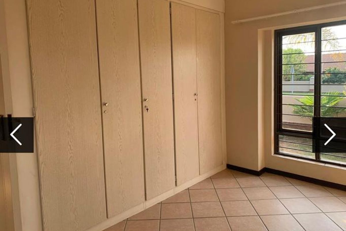 A clean, empty bedroom featuring neutral tiled flooring and extensive built-in cupboard storage with natural light from a window fitted with security bars.