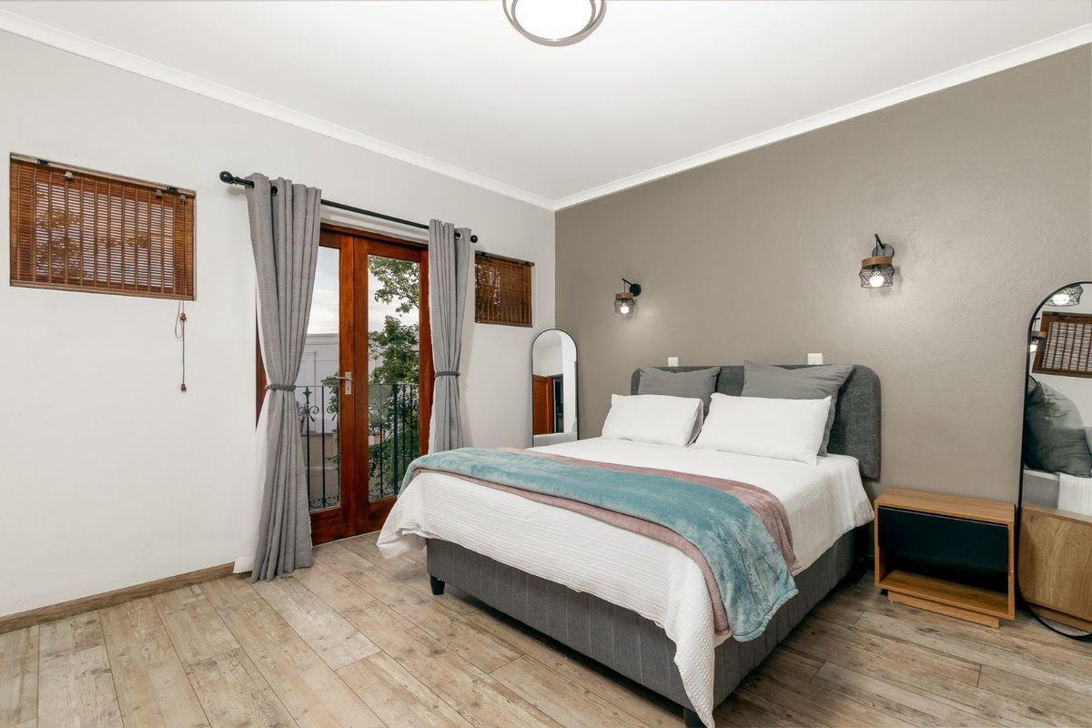 A well-maintained bedroom featuring timber-look flooring, a feature wall, wall-mounted lighting, and direct access to a private balcony through timber french doors.