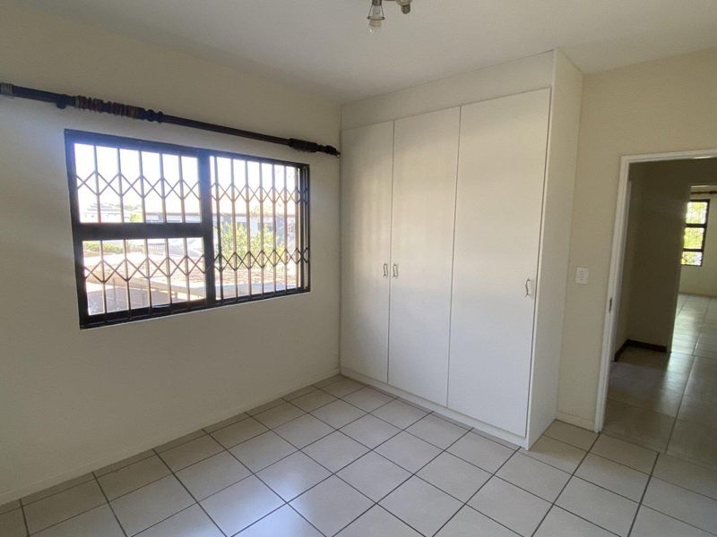 A clean, empty bedroom featuring tiled flooring, built-in cupboards, and a window fitted with a security gate.