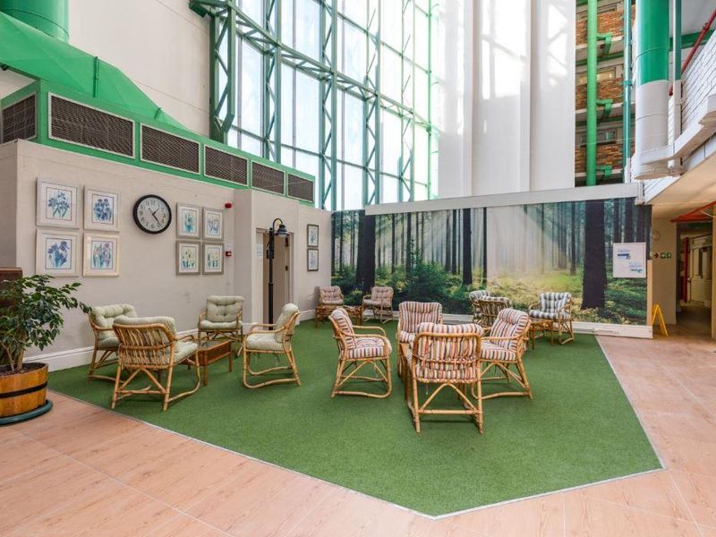 Spacious indoor communal area featuring high ceilings, full-height glass windows, exposed industrial ducting, and a dedicated artificial turf seating zone.