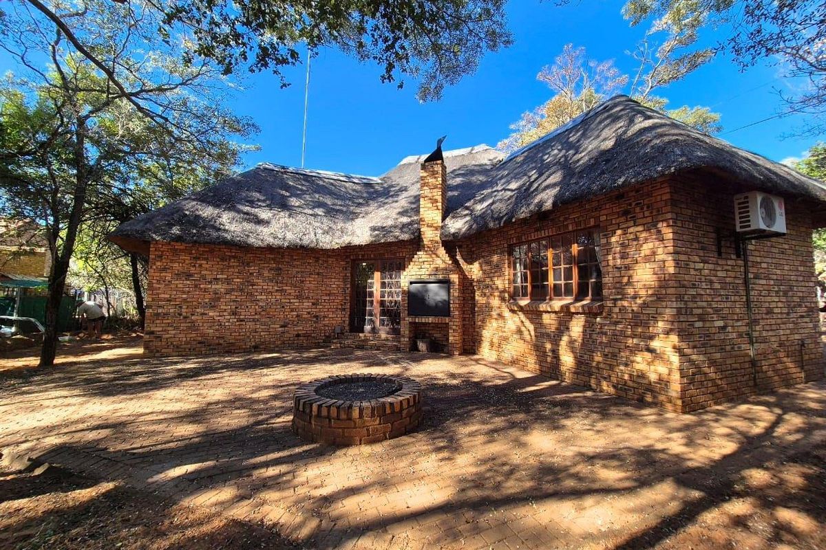 A rustic brick house featuring a traditional thatched roof, an outdoor brick fireplace, and a paved patio area set within a natural garden setting.