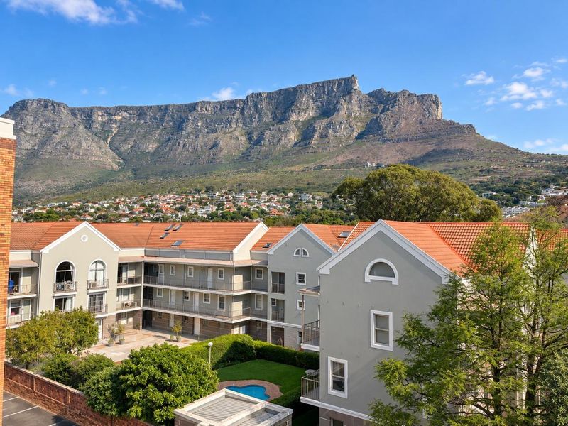 Aerial view of an apartment complex featuring terracotta roofs, balconies, and a communal garden with a swimming pool, set against a mountain backdrop.