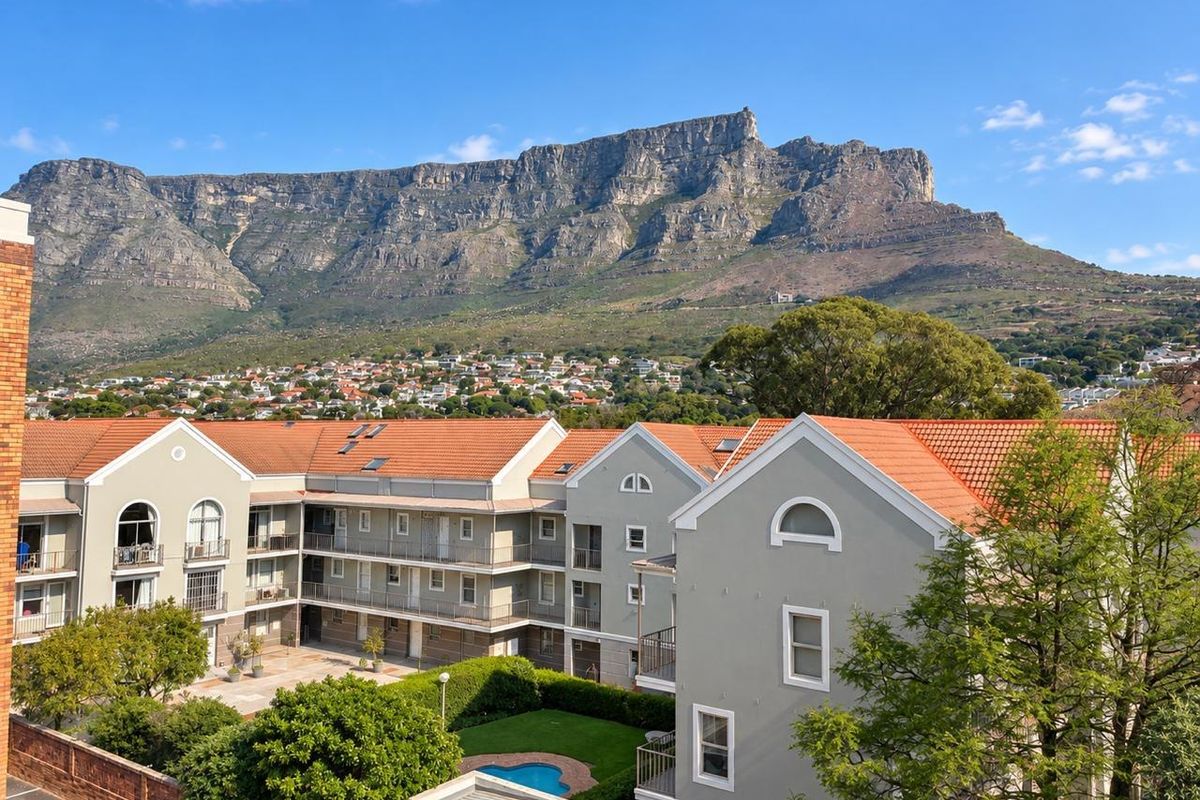 Aerial view of an apartment complex featuring terracotta roofs, balconies, and a communal garden with a swimming pool, set against a mountain backdrop.