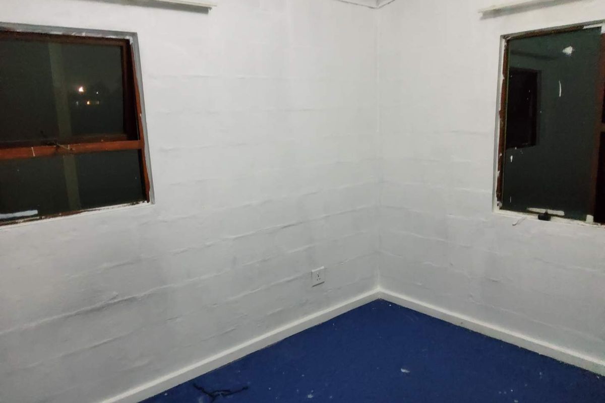 An empty bedroom featuring white painted block walls, blue carpeting, and two windows with wooden frames.