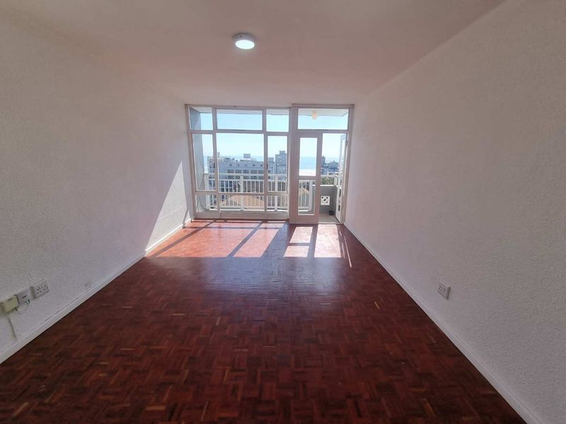 An empty living room featuring classic parquet flooring, a large glass door leading to a balcony, and ample natural light.