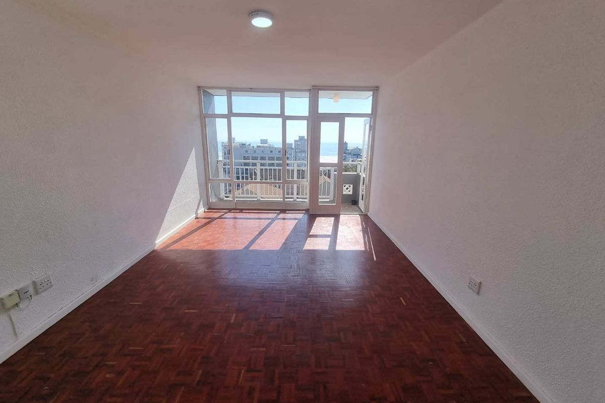 An empty living room featuring classic parquet flooring, a large glass door leading to a balcony, and ample natural light.