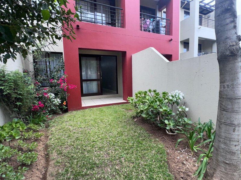 A private rear garden area featuring a small lawn, landscaped borders, and a tiled patio leading to a sliding glass door entrance.