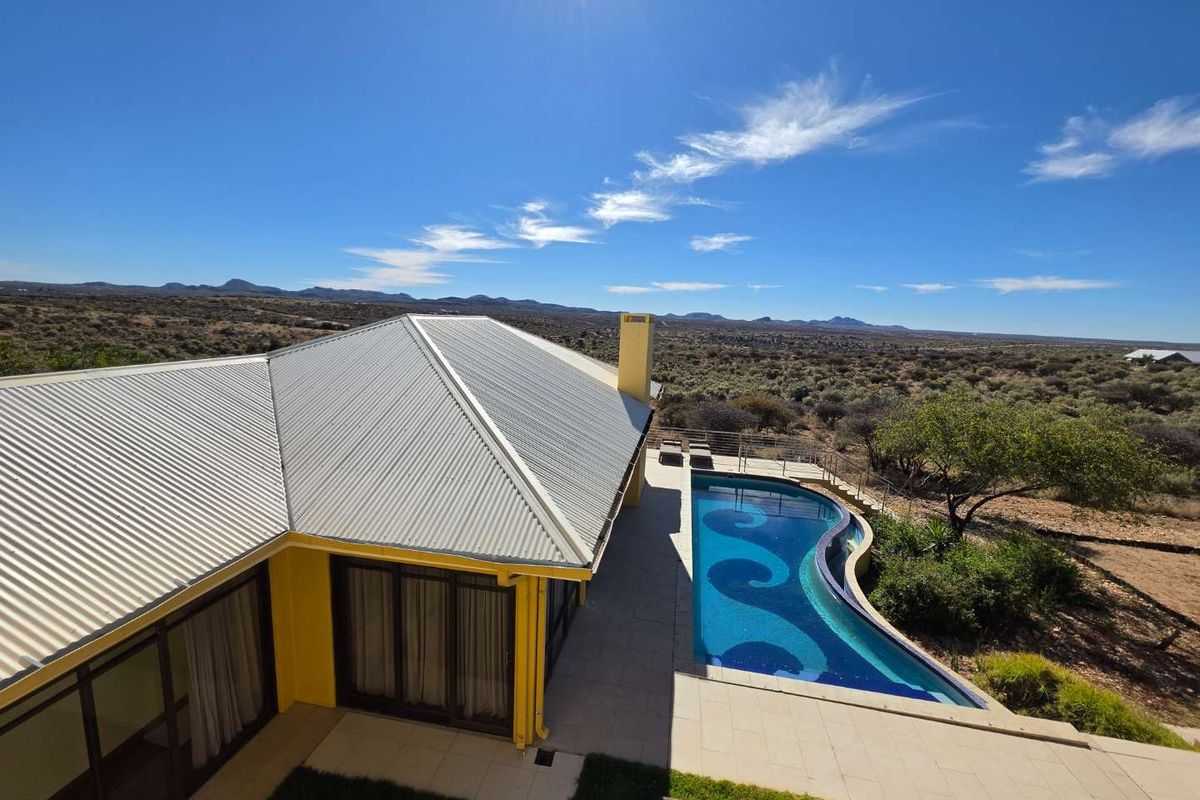 Aerial view of a residential property featuring a curved swimming pool, paved patio area, and metal roof, set against a vast landscape.