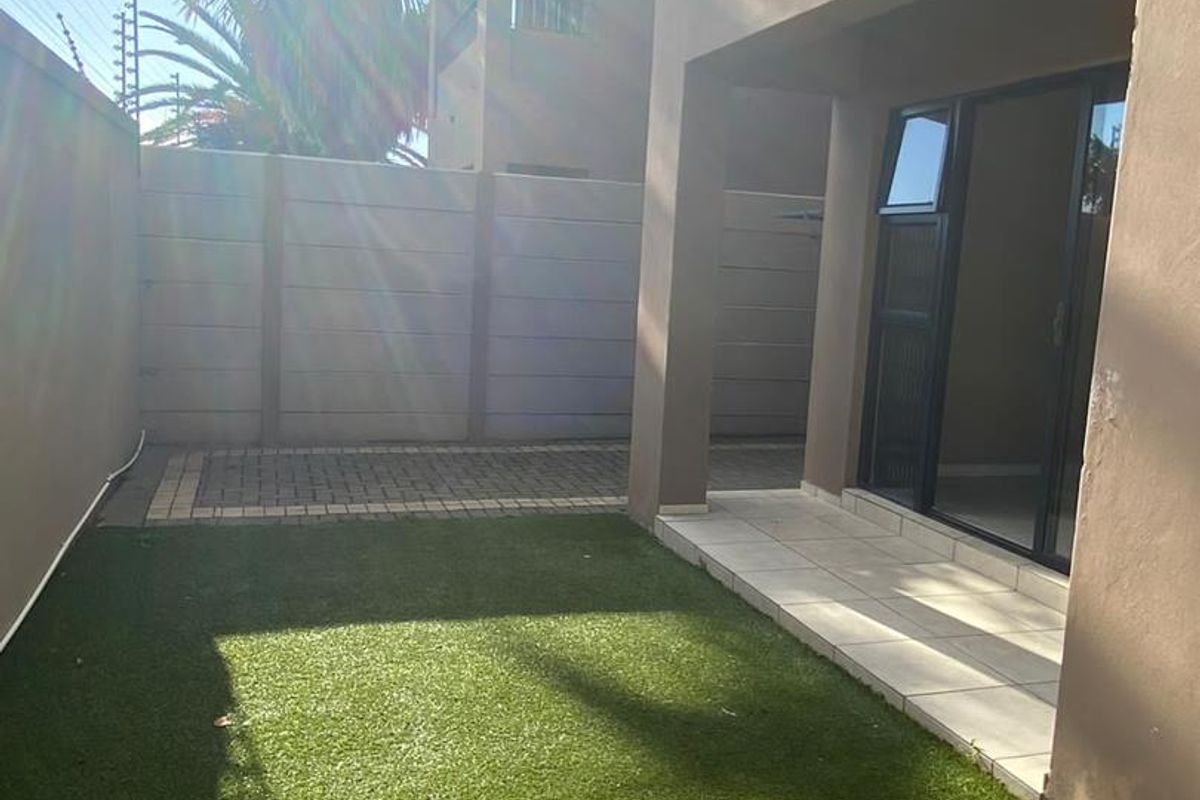 A private rear courtyard featuring a paved patio, artificial lawn, and secure boundary walling, accessible via a sliding glass door.