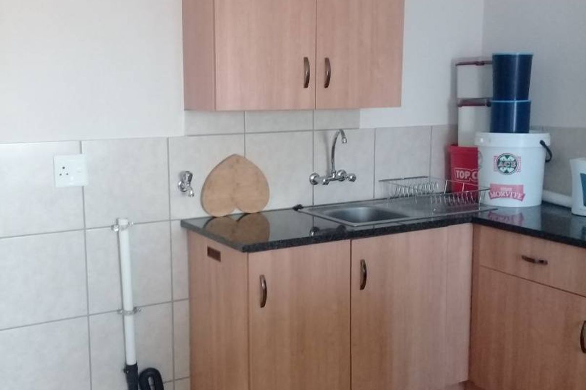A functional kitchen area featuring laminate cabinetry, granite countertops, a stainless steel sink, and tiled splashbacks with visible plumbing connections.