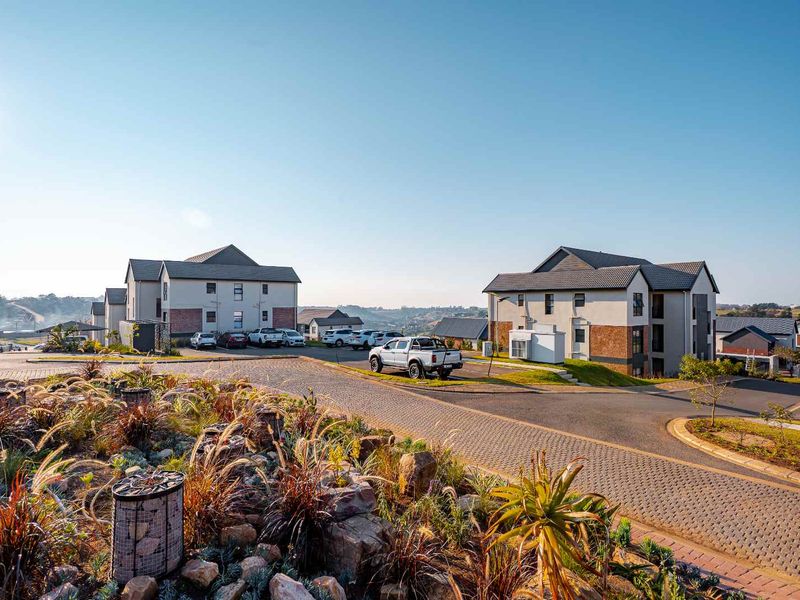 A wide-angle view of a modern apartment complex featuring paved driveways, landscaped gardens, and multi-story residential buildings with tiled roofs.