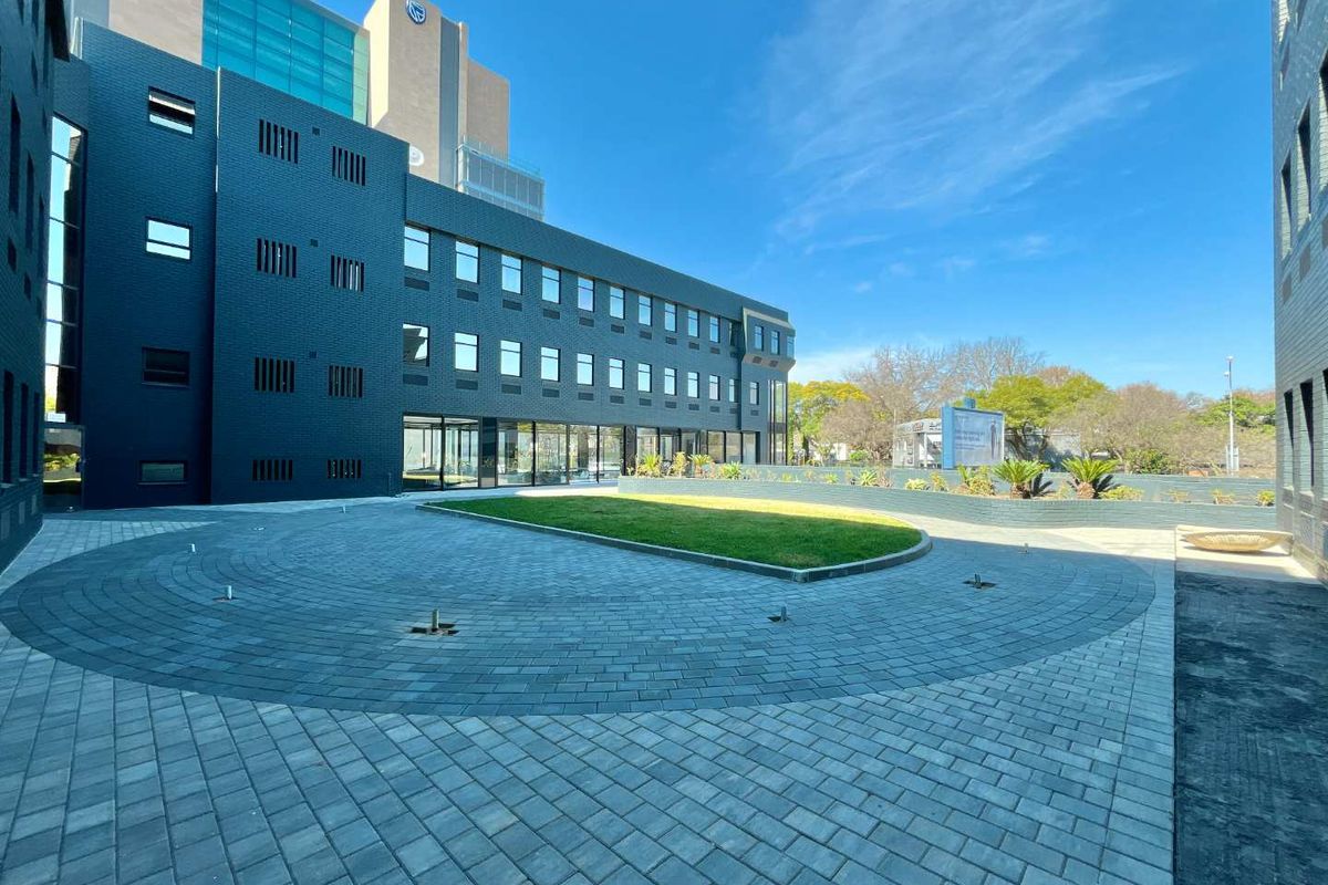 A modern office building exterior featuring a paved courtyard, landscaped lawn, and a combination of dark brick and glass facade elements.