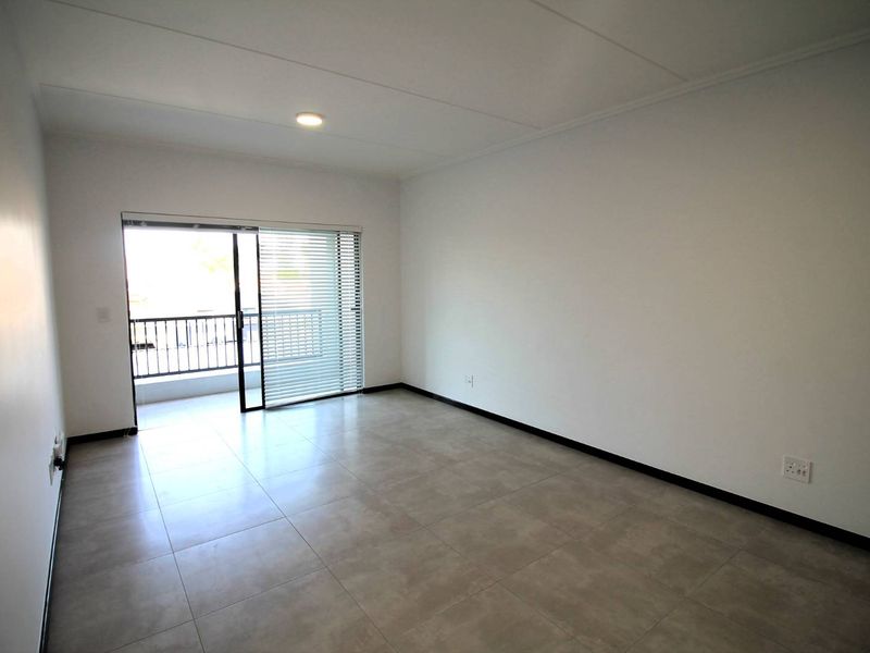 A bright, empty living room featuring neutral tiled flooring, clean white walls, and a sliding glass door providing access to a balcony.
