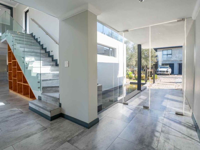 Modern entrance hall featuring a glass staircase, built-in shelving, tiled flooring, and a seamless glass pivot door opening to an outdoor water feature.