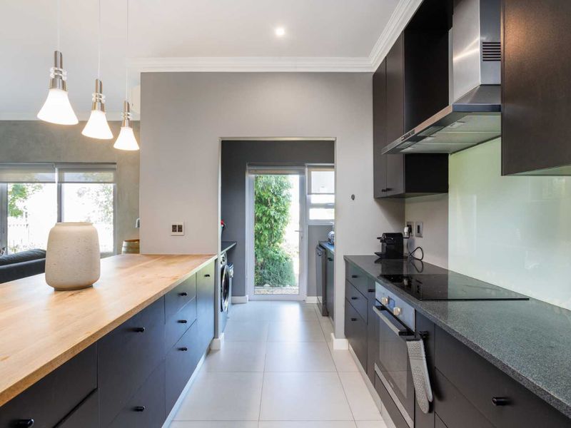 Modern kitchen featuring dark cabinetry, stone countertops, a timber breakfast bar, integrated appliances, and a glass splashback, leading to a separate laundry area.