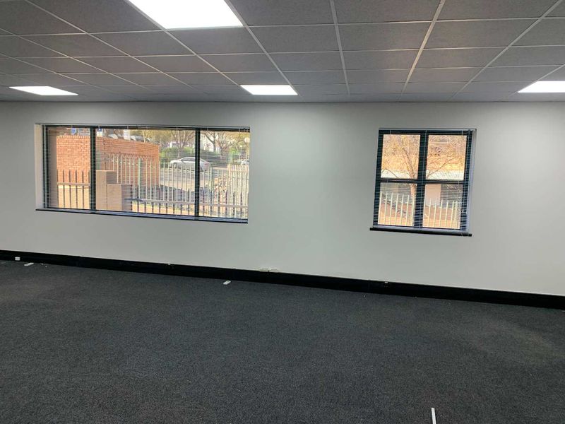 A bright, open-plan office space featuring a suspended ceiling with recessed LED lighting, carpeted flooring, and large windows with fitted venetian blinds.