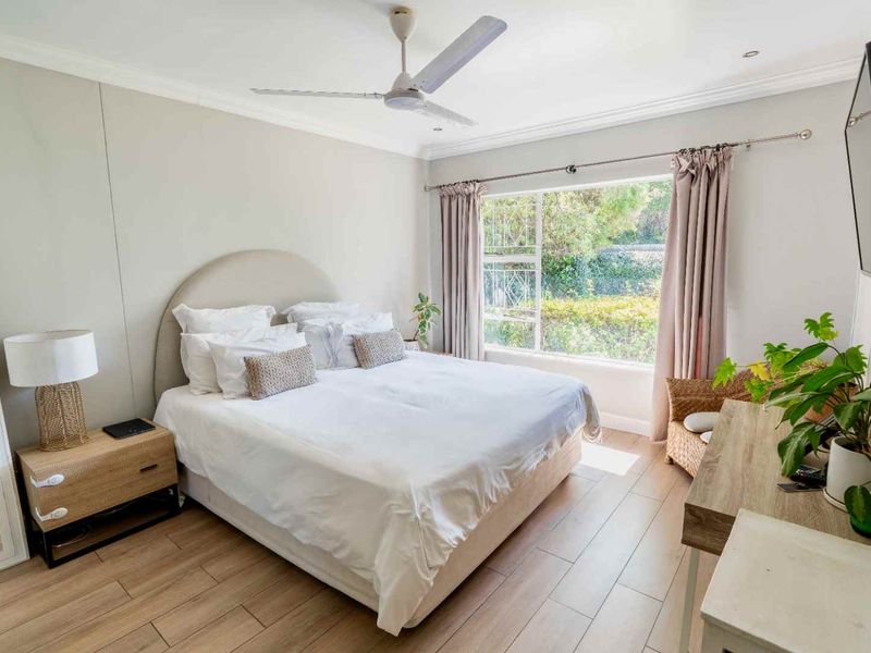 A bright and well-maintained bedroom featuring laminate flooring, a ceiling fan, and a large window providing natural light and garden views.
