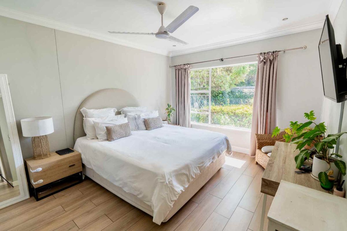 A bright and well-maintained bedroom featuring laminate flooring, a ceiling fan, and a large window providing natural light and garden views.