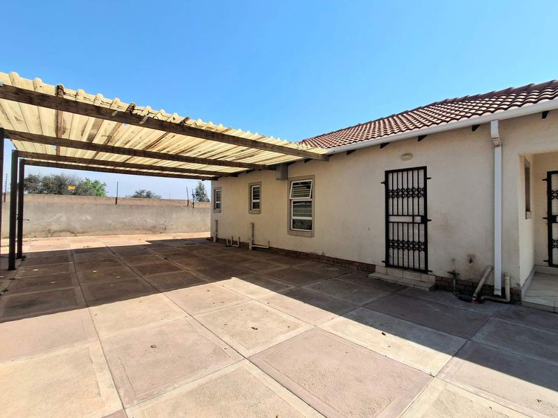 Rear exterior view featuring a paved patio area, a covered carport structure, and secure perimeter walling with electric fencing.