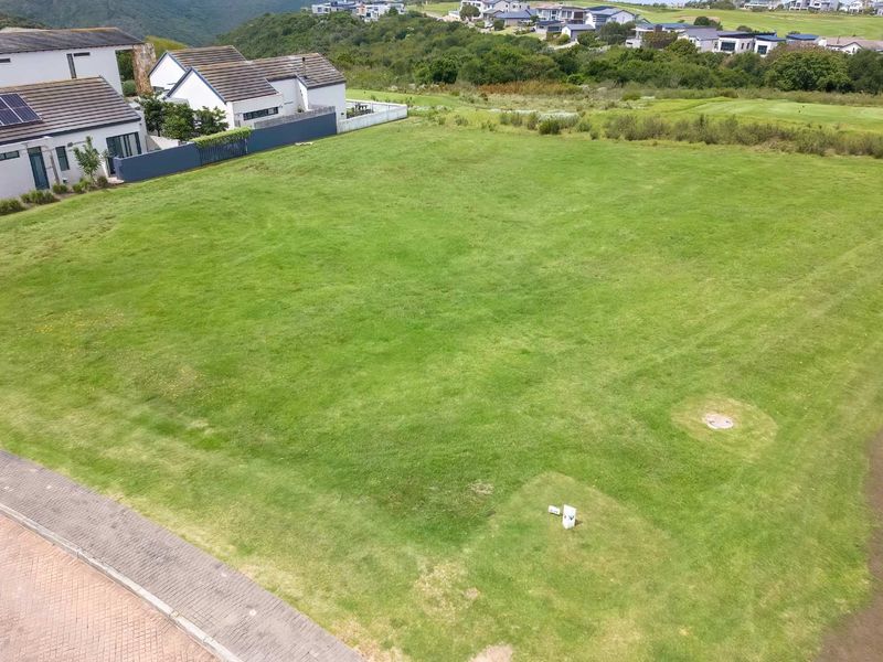 An aerial view of a vacant residential plot featuring a level grassed surface, utility access points, and adjacent paved road access in a developed estate.