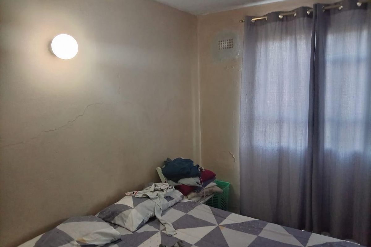 A bedroom featuring a wall-mounted light fixture and ventilation grille, showing signs of wear including cracked plaster and peeling paint.