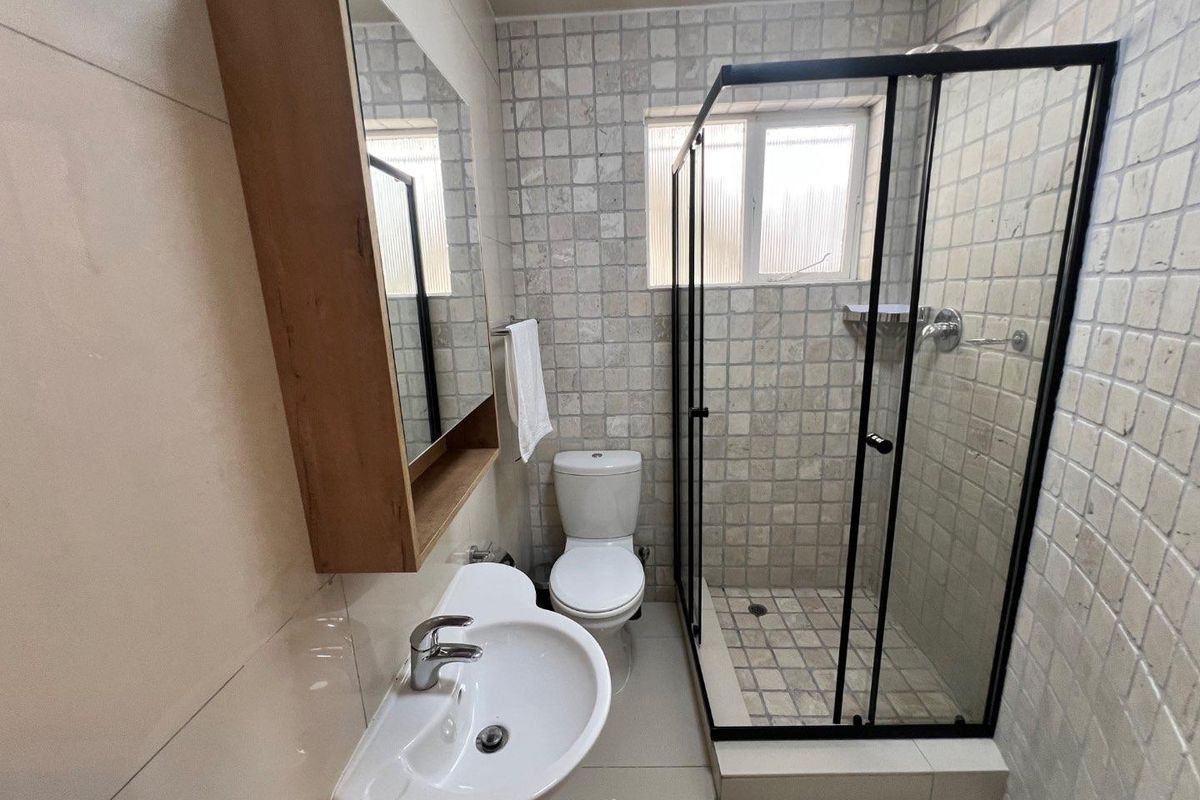 A clean, functional bathroom featuring a glass-enclosed shower, pedestal basin, and wall-mounted mirror cabinet with neutral tiled finishes.