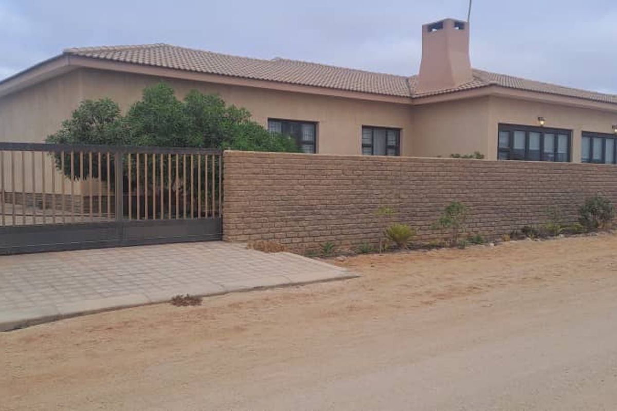 Front view of a single-story house featuring a perimeter wall, sliding gate, paved driveway, and a prominent chimney.