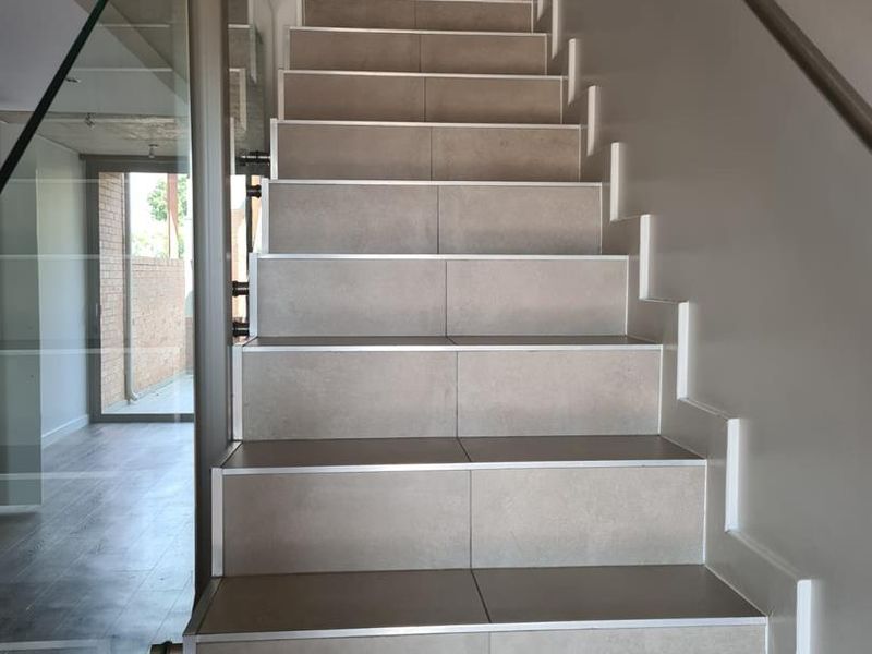 Modern staircase featuring tiled steps with metal nosing, complemented by a sleek glass balustrade and a wall-mounted metal handrail.