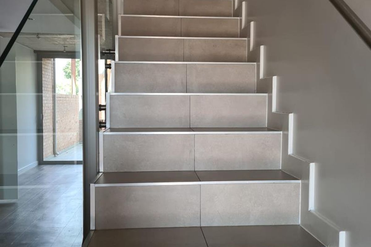 Modern staircase featuring tiled steps with metal nosing, complemented by a sleek glass balustrade and a wall-mounted metal handrail.