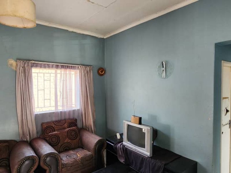 A living room area featuring blue walls, a window with security bars, and a ceiling light fixture, showing signs of wear and surface damage.