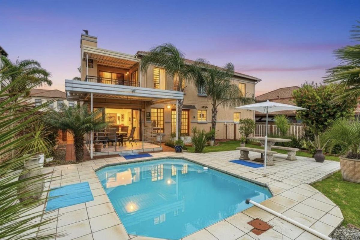 Rear view of the house featuring a swimming pool, paved pool deck, covered patio area, and an upper-level balcony.