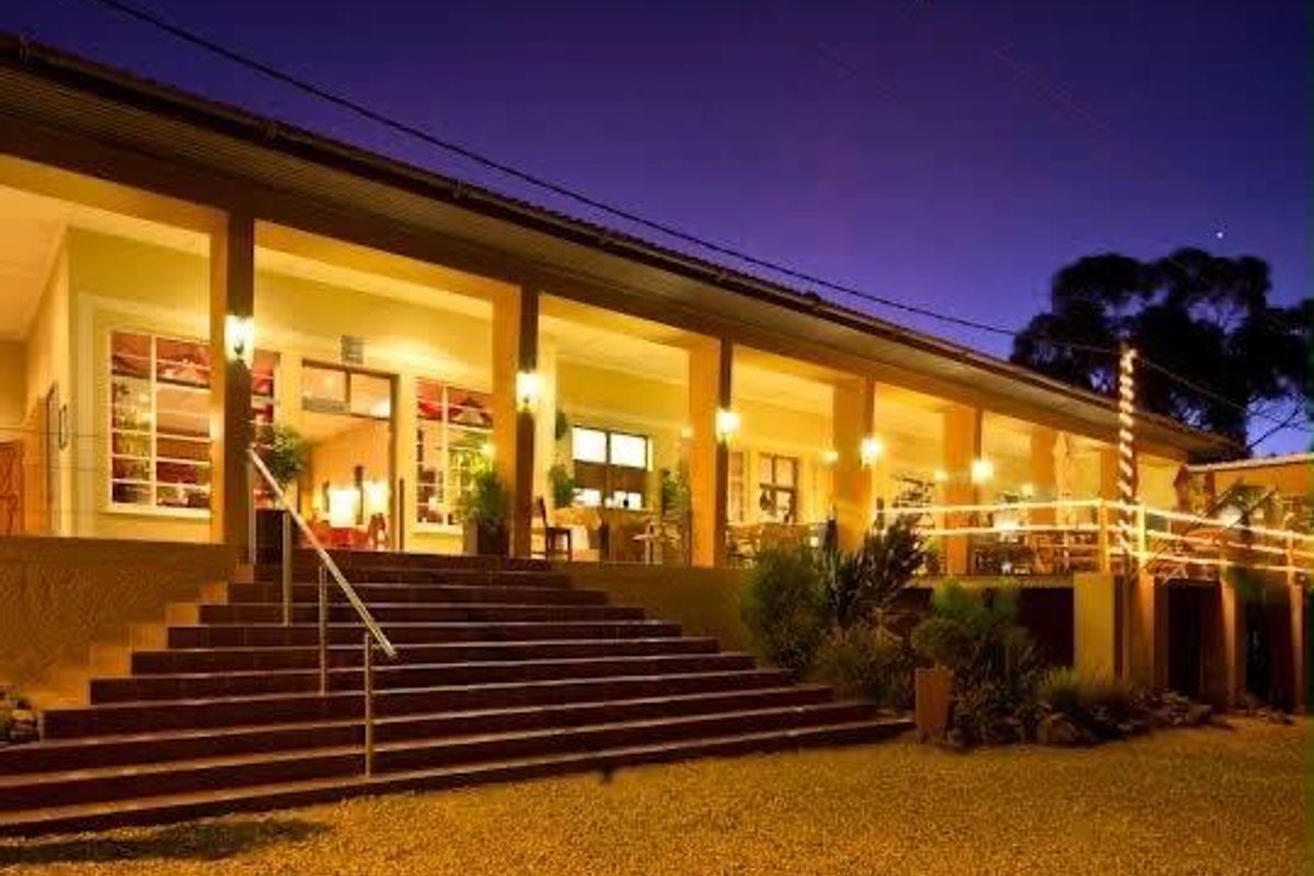 A well-lit rear exterior view of the hotel featuring a spacious covered veranda, concrete staircase with metal handrails, and timber balustrades.