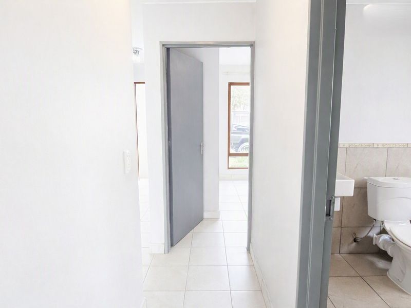 A bright, neutral hallway connecting to a bathroom and adjacent rooms, featuring clean tiled flooring and simple, modern finishes throughout.