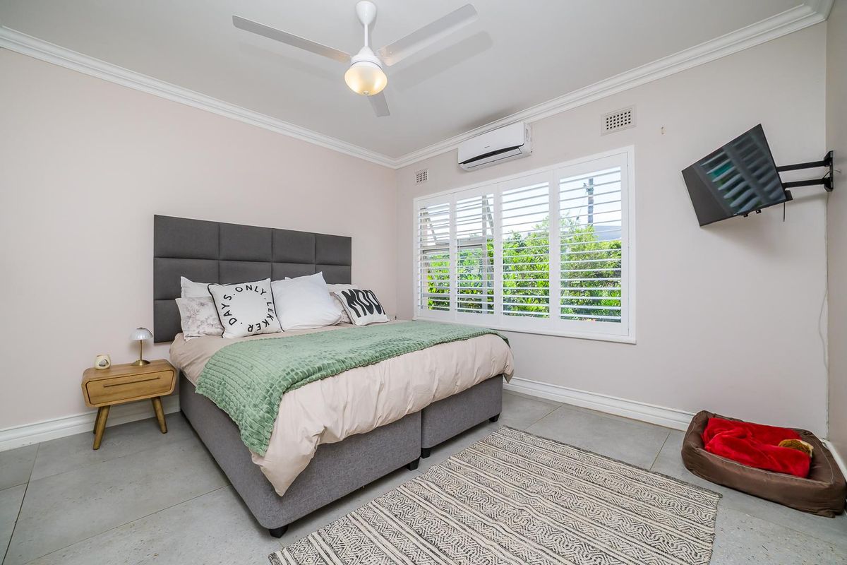 A well-maintained bedroom featuring tiled flooring, plantation shutters, a ceiling fan, and a wall-mounted air conditioning unit.