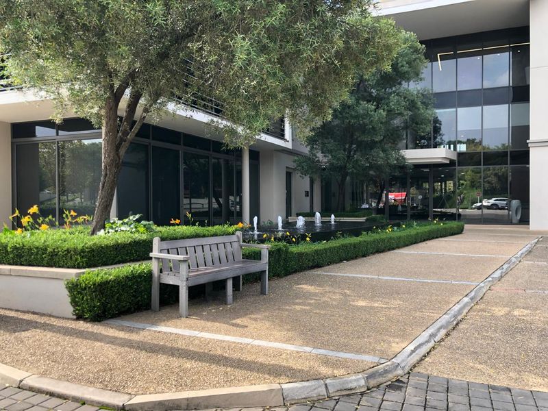 Modern office building exterior featuring a landscaped garden, water feature, and paved pedestrian walkway leading to a glass-fronted entrance.