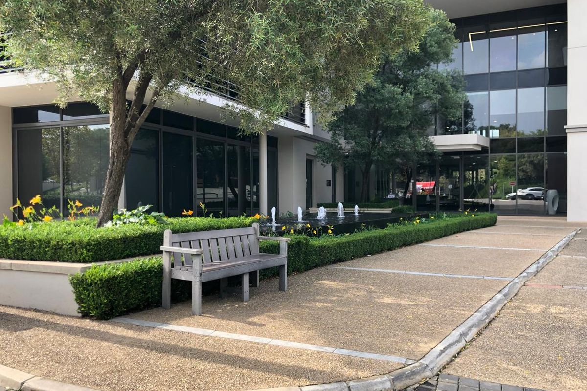 Modern office building exterior featuring a landscaped garden, water feature, and paved pedestrian walkway leading to a glass-fronted entrance.