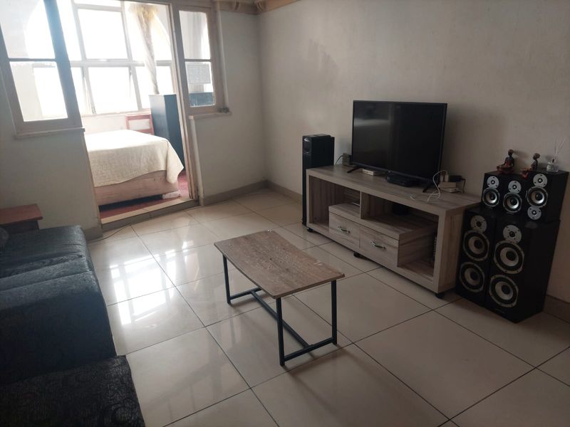 A living room featuring tiled flooring and a large window, providing access to an adjacent bedroom area.