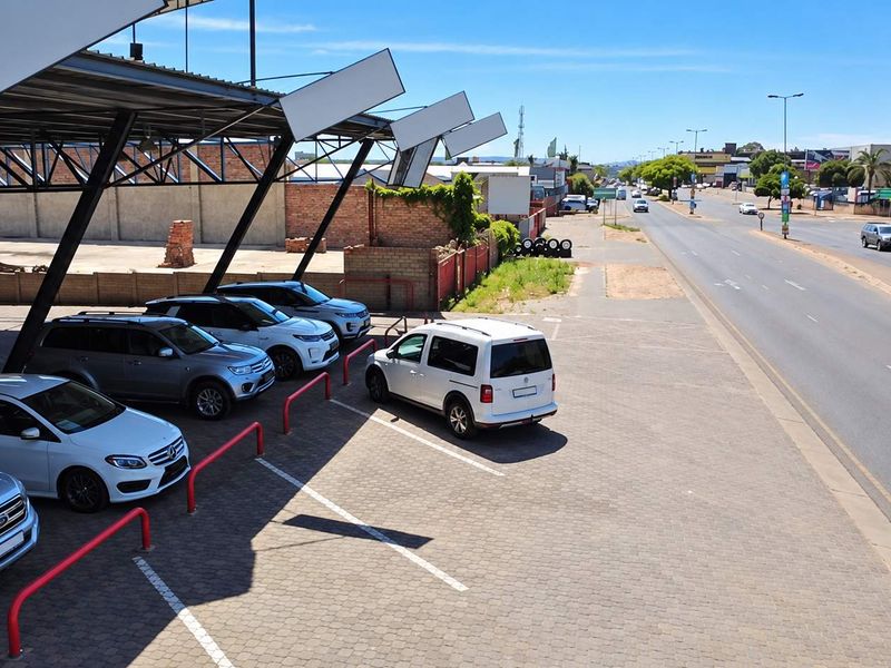 Exterior view of a retail property featuring a paved parking area with a metal carport structure and brick boundary wall along a main road.