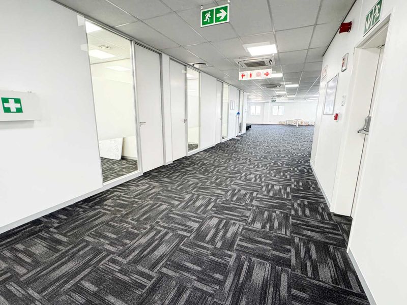 A clean, modern office hallway featuring carpeted flooring, suspended ceilings with recessed lighting, and multiple glass-partitioned office entrances.