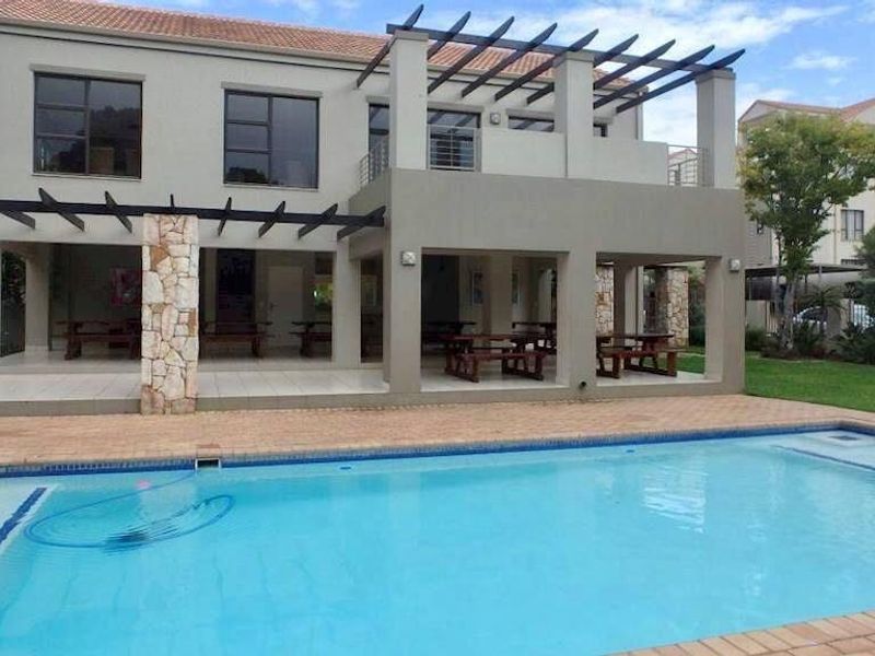 A well-maintained apartment complex pool area featuring a large swimming pool, covered patio with stone-clad pillars, and an upper-level balcony with pergola.