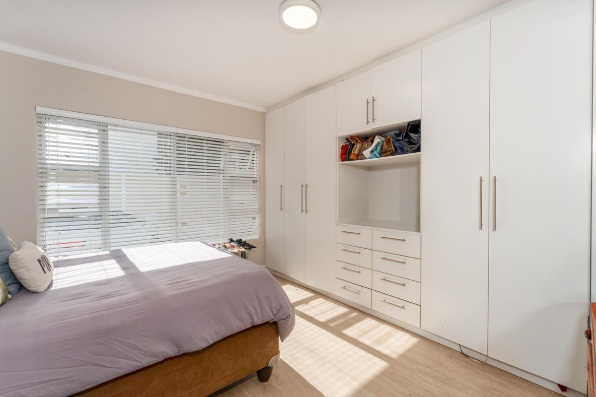 A bright bedroom featuring extensive built-in white cabinetry with integrated drawers and shelving, complemented by light wood-look flooring and venetian blinds.