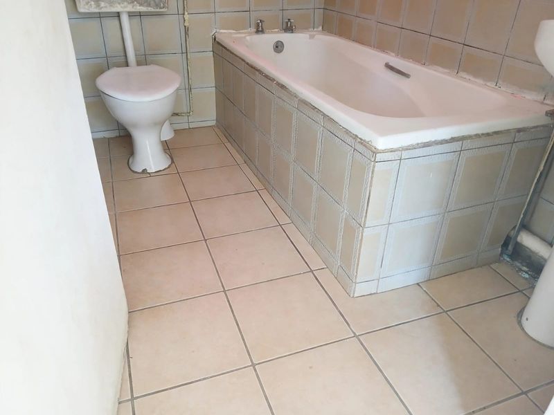 A dated bathroom featuring a built-in bathtub, pedestal basin, and toilet, finished with original ceramic wall and floor tiling.