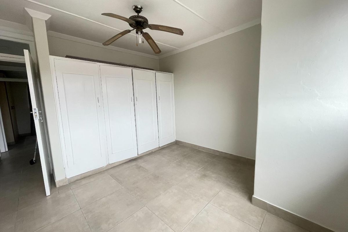 A bright, empty bedroom featuring neutral tiled flooring, built-in cupboards, and a ceiling fan, offering a clean canvas for personalization.