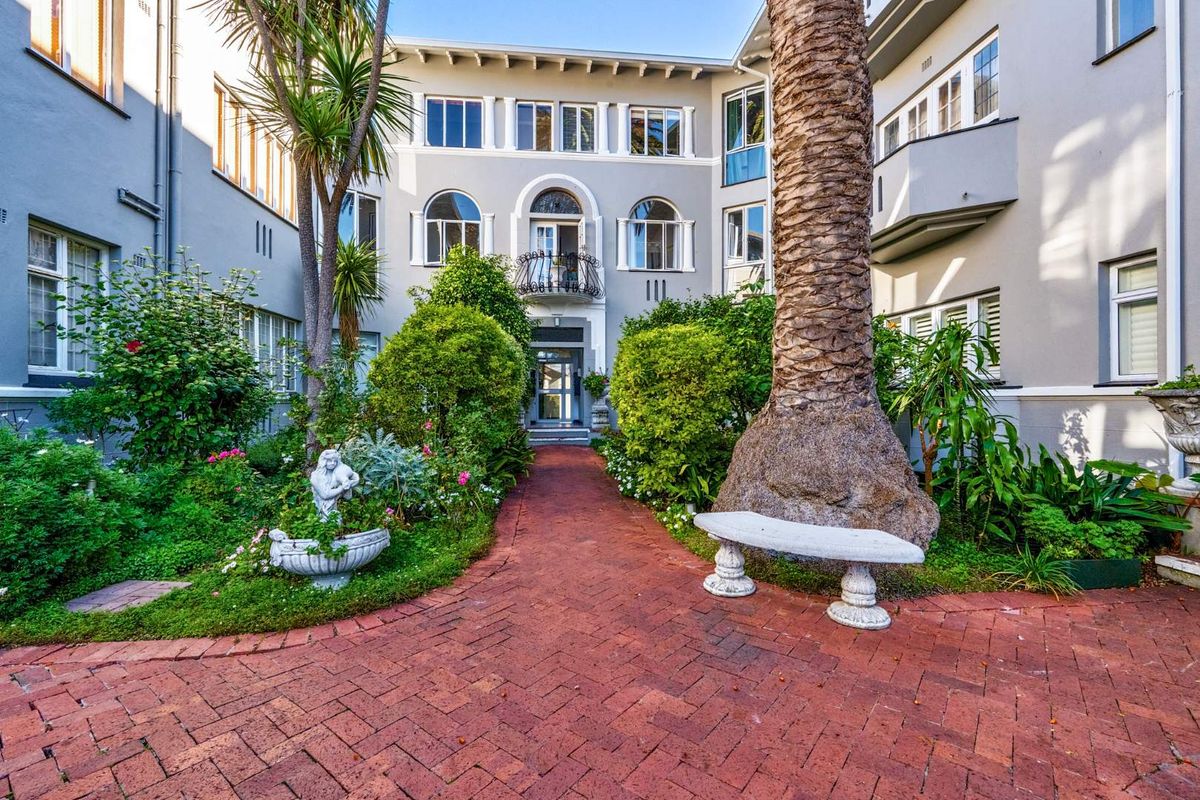 A charming courtyard garden featuring a brick-paved pathway leading to a multi-story apartment building with classic architectural details and arched windows.