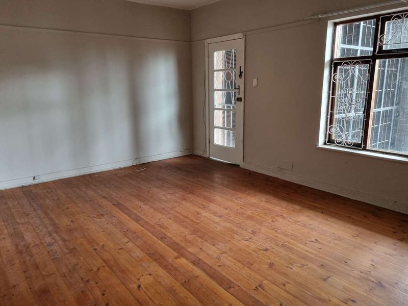 An empty living room featuring original hardwood flooring, a glass-paneled door, and large windows with security bars, offering a blank canvas for renovation.